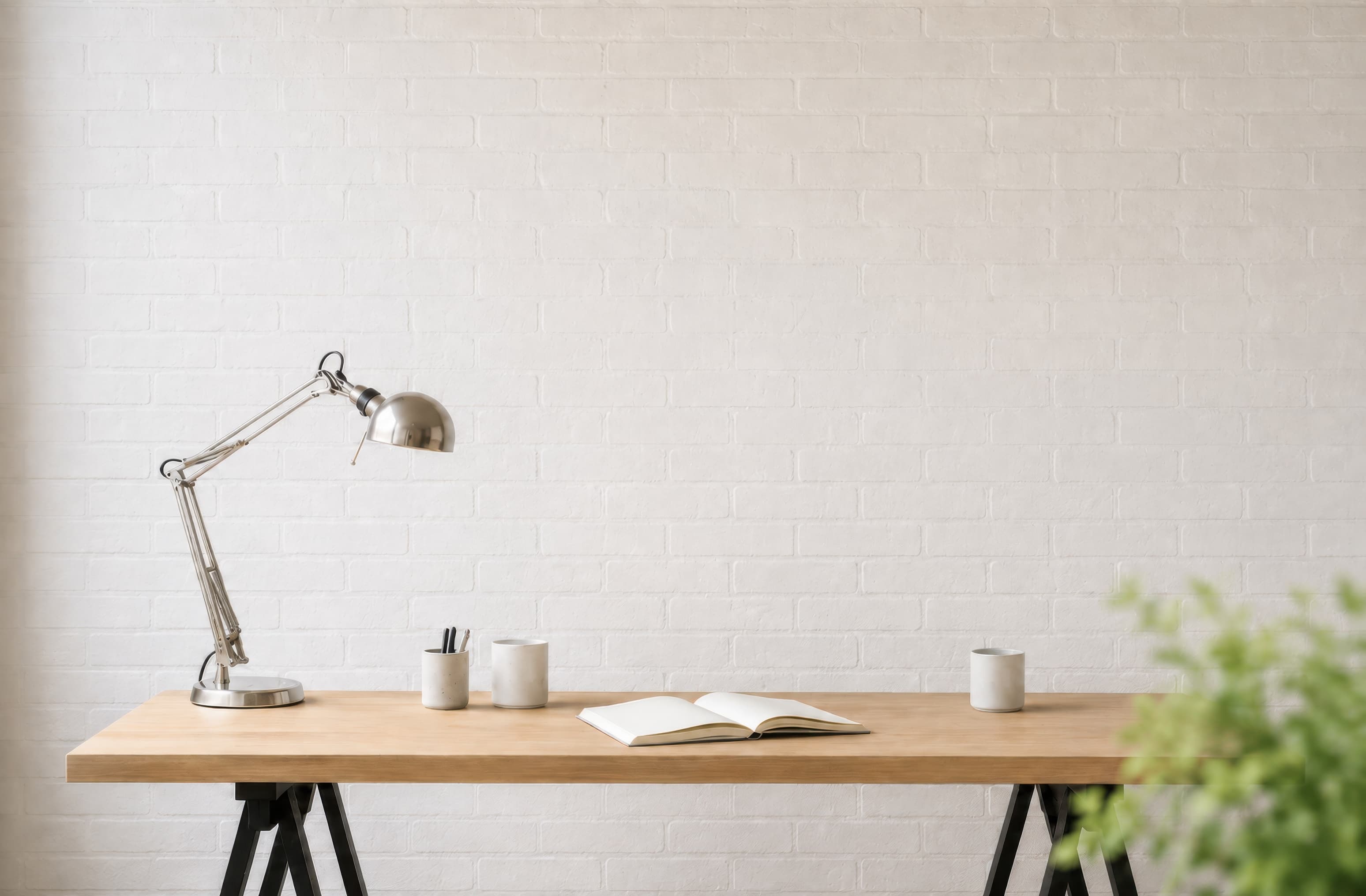 Minimal white brick office wall above a pale wood desk with a chrome lamp, cups, notebook, and blurred green plant., ranked 6 among office mockups