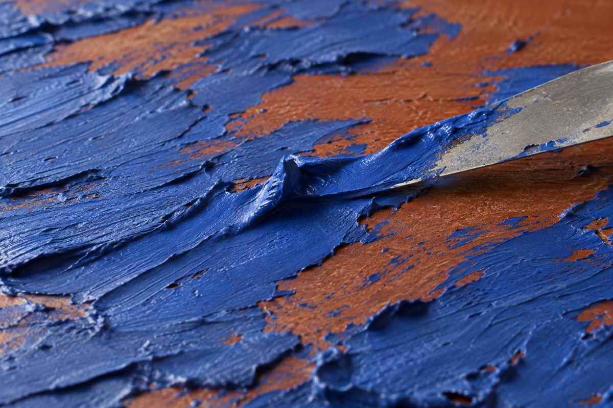 Close process crop of a painting knife midway through a low-angle drag, leaving blue acrylic ridges over a dry sienna ground